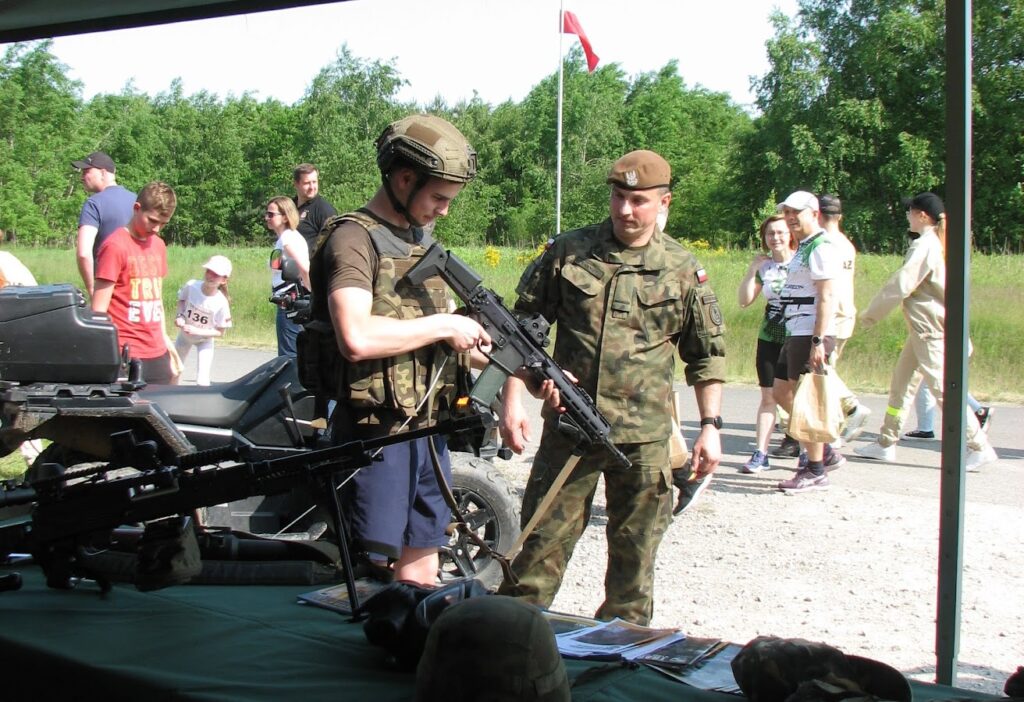 Na zdjęciu w tel przechadzający się biegacze, na froncie żołnierz tłumaczący obsługę broni. 