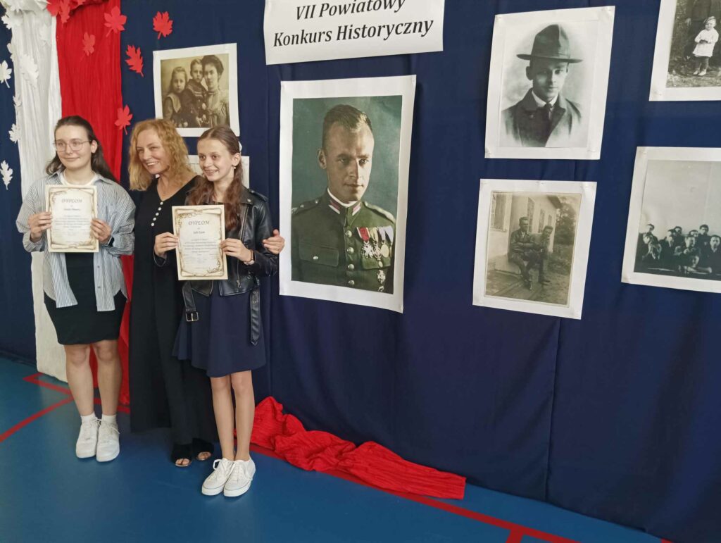 Na tle dużej tablicy z wizerunkami Witolda Pileckiego stoją laureatki konkursu z członkinią komisji konkursowej. 